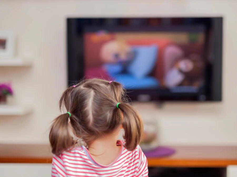 Ein Kind mit Zöpfen sehen wir von hinten. Es schaut Kabelfernsehen.